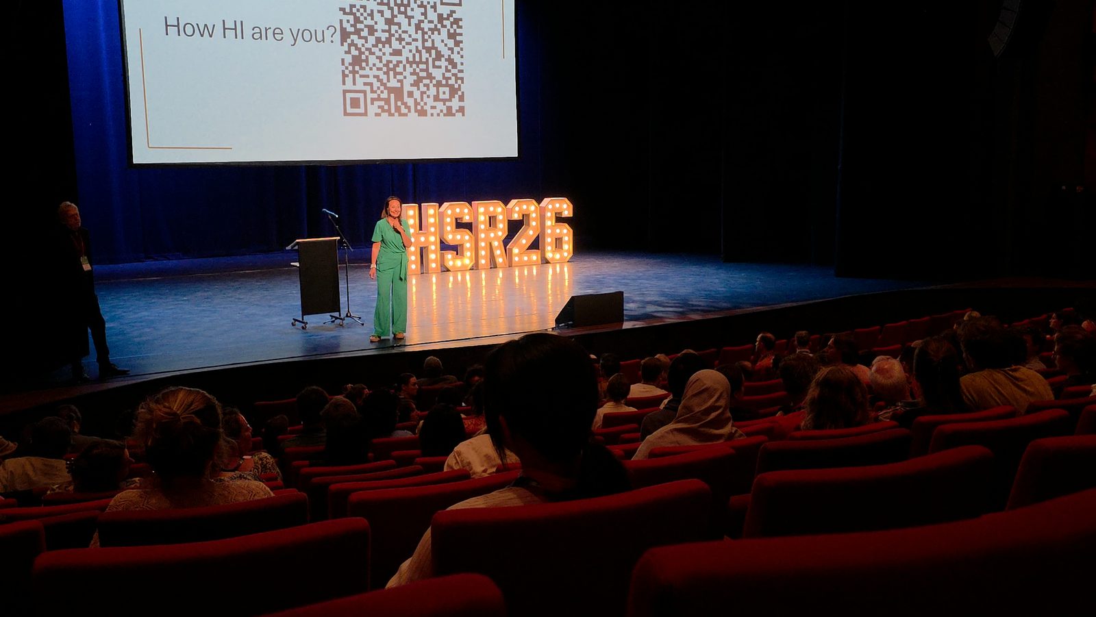 Debby Mureau op het podium van een theater, met op het scherm How HI are you?