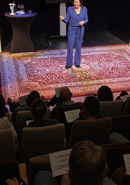 Debby Mureau op een podium met een groot tapijt, het publiek kijkt naar haar